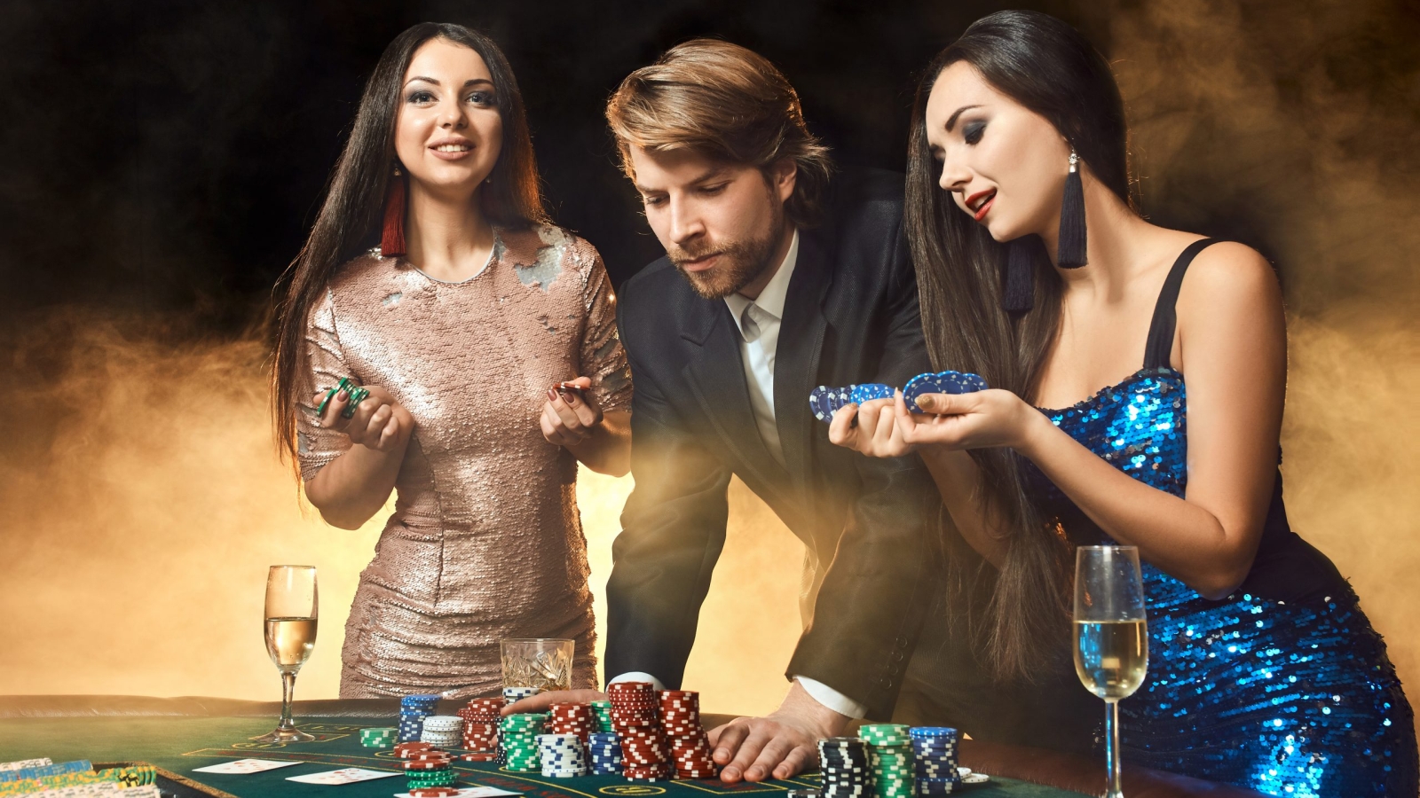 Two beautiful women and young man play on poker table in casino, focus on man and brunette. Passion, cards, chips, alcohol, dice, gambling, casino - it is entertainment. Dangerous fun card game for money. Smoke background.